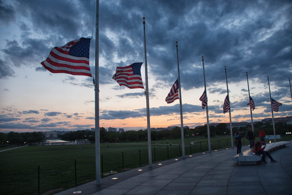 political violence is on the rise in America. Image shows flags flying at half-mast at the base of the Washington Monument, at sunset.